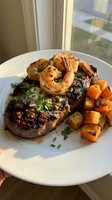 Grilled steak topped with seared shrimp, roasted sweet potato cubes, and fresh herbs on white plate by window.