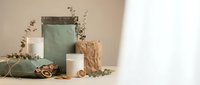 Minimalist gift wrapping setup with sage green and kraft paper bags, white candle, eucalyptus branches and golden scissors on neutral beige background.