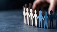 Paper cutout figures holding hands in curved line, white and blue silhouettes symbolizing unity, teamwork and diversity on dark background with selective focus.