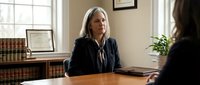 Mature female attorney with gray hair wearing dark blazer conducts client consultation in professional law office with wooden desk and bookshelf.