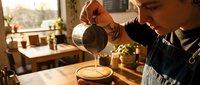 Professional barista pouring steamed milk creating latte art in coffee cup at modern cafe counter during golden hour morning light.