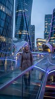 Young blonde businesswoman in beige coat standing on glass bridge in modern financial district at dusk with illuminated skyscrapers and city lights.