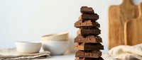 Stacked dark chocolate pieces arranged in tower formation with blurred kitchen bowls and wooden utensils in background, suitable for food photography and culinary content.