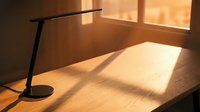 Minimalist workspace with black desk lamp casting shadows on wooden table surface, warm sunlight streaming through window creating geometric light patterns.