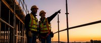 Two construction workers in safety vests and hard hats discussing project plans on building site scaffolding during golden hour sunset.