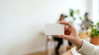 Close-up of hand holding blank white business card with blurred office background and potted plants, suitable for branding and corporate identity mockups.