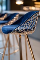 Modern bar stool with navy blue velvet seat and intricate golden geometric pattern backrest against blurred bokeh background in luxury interior setting.