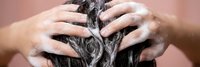 Woman washing long dark hair with shampoo foam in shower, hands massaging scalp for healthy hair care routine and personal hygiene maintenance.