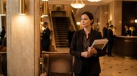 Professional businesswoman in elegant black suit holding documents in luxurious hotel lobby with marble columns and warm lighting for corporate hospitality concepts.