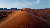 Rippled sand dunes stretch across vast desert landscape at sunset, dramatic orange ridges contrast against blue mountains and sky, natural patterns in sand formation.