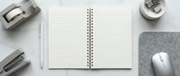 Clean minimalist workspace with open spiral notebook, computer mouse, stapler and office supplies on white wooden desk for productivity and organization.