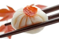 Steamed Asian dumpling topped with fresh salmon held by wooden chopsticks against blurred red background, close-up view of traditional dim sum cuisine.