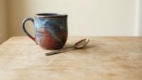Handcrafted ceramic coffee mug with blue and brown glazed finish sits on wooden table with vintage spoon, perfect for cozy morning rituals and cafe decor.