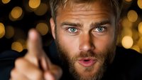 Confident bearded man with blue eyes pointing finger at camera against blurred golden bokeh lights background, expressing positive attitude and friendly approach.