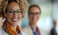 Mixed race businesswoman with curly blonde hair and glasses smiling confidently in office setting, wearing colorful scarf. Professional corporate environment with coworker in background.