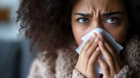 Young African American woman with curly hair blowing nose in tissue paper, showing symptoms of seasonal flu, cold or allergies while looking concerned.