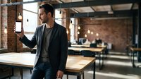 Young professional businessman using smartphone in modern industrial office space with exposed brick walls and pendant lighting for business communication.