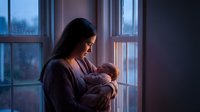 Young mother holding newborn baby by window at twilight, tender maternal bonding moment in cozy home interior for parenting and family concepts.