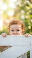 Adorable curly-haired toddler peeking over white wooden fence with bright smile in sunny garden setting for family and childhood concepts.