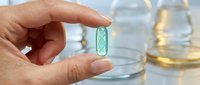 Hand holding green herbal supplement capsule with glass of water in background for health and wellness concept.