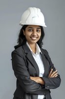 Young Indian female engineer in business suit and white hard hat smiling confidently with arms crossed against gray background. Professional portrait for construction industry.
