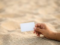Hand holding blank white card on sandy beach surface, suitable for business cards, travel promotions, vacation marketing, and summer campaigns.