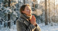 Young woman with closed eyes in prayer pose enjoying peaceful winter morning with falling snow and warm sunlight filtering through frost-covered trees.