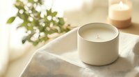 Minimalist white scented candle in ceramic holder on soft textile with green plant and glowing candle in background, creating cozy atmosphere.