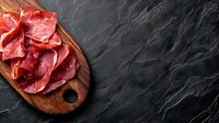 Thinly sliced cured meat arranged on wooden cutting board against dark slate background. Traditional Italian antipasto or charcuterie presentation.