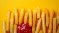 Crispy golden french fries arranged in row with red ketchup splash on vibrant yellow background. Modern food photography for restaurant menu or advertising.