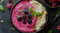 Vibrant pink smoothie bowl with creamy white swirl, topped with fresh blackberries, raspberries, chia seeds and mint leaves on dark background.