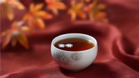 Hot tea served in traditional Asian ceramic cup with floral pattern on burgundy silk fabric background with orange autumn maple leaves, cozy fall atmosphere.