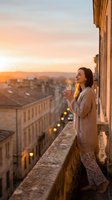 Woman in beige cardigan holding coffee cup on historic building balcony overlooking cityscape at golden hour sunset, urban travel and lifestyle moment.