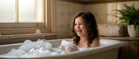 Young girl with curly hair enjoying relaxing bubble bath in modern bathroom with natural light and plants for wellness and self-care concepts.