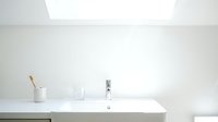 Minimalist bathroom sink with white countertop, chrome faucet, and organized toiletries in ceramic holder under bright skylight for modern home design.