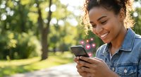 Young African American woman smiling while using smartphone outdoors in sunny park with heart emojis floating from device suggesting social media love.