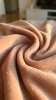 Soft beige fleece blanket with swirled folds creating cozy texture in warm sunlight on living room sofa for comfort and relaxation at home.
