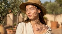 Woman with long dark hair and olive skin tone wearing a tan straw hat and cream linen shirt, standing in a Mediterranean garden with olive trees and terracotta pots during golden hour sunlight.