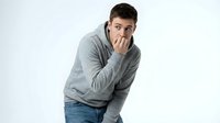 Young Caucasian man in gray hoodie and jeans biting fingernails, expressing anxiety, stress, or nervous habit against white background.