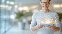 Caucasian businessman in glasses holding digital tablet with glowing magical light effect emerging from screen, modern office interior with bokeh background.