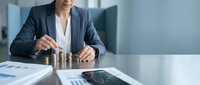 Professional businesswoman stacking golden coins at modern office desk with financial charts and tablet displaying growth data for investment planning.