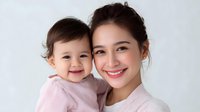 Young Asian mother and baby daughter smiling together against white background, both wearing pink, expressing joy and family bonding in natural light portrait.