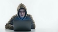 Hooded hacker in dark clothing working on laptop computer in dimly lit environment for cybersecurity and digital crime concepts.