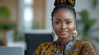 Young African businesswoman with braided hair and gold earrings wearing traditional print ankara blouse smiles confidently in modern office setting, representing success.