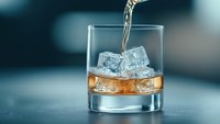 Pouring amber liquid into rocks glass with clear ice cubes, close-up shot against blurred blue background showing drink preparation and mixology.