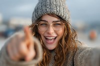 Cheerful young woman with glasses and knit hat pointing at camera, expressing joy with bright smile. Natural outdoor setting with blurred background.