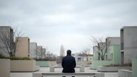 Man in black jacket sitting on bench overlooking urban cityscape with modern buildings and bare trees during overcast day, contemplative mood for lifestyle and wellness content.
