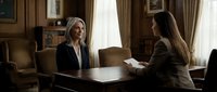 Two professional Caucasian women in formal attire having a serious business meeting in an elegant wood-paneled office with bookshelves.