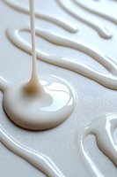 Flowing white liquid drops and streams creating abstract patterns on light surface, macro photography showing viscous texture and glossy finish.