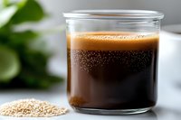 Fresh brewed coffee in glass cup with raw coffee beans scattered on marble surface, selective focus on dark liquid with crema foam on top.
